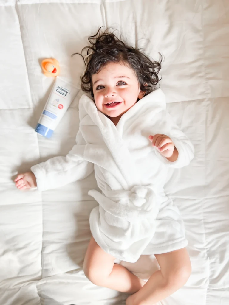 A smiling toddler in a white robe with a Happy Cappy Moisturizing Cream tube beside them