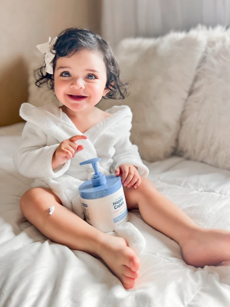 A smiling toddler in a white robe holding a Happy Cappy Moisturizing Cream pump jar