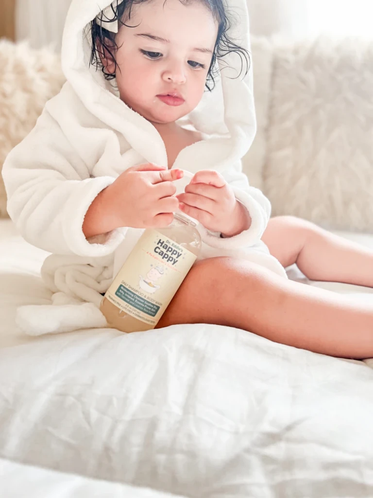 A toddler in a white hooded robe holding a Happy Cappy Daily Shampoo & Body Wash bottle