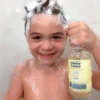 Smiling boy covered in lather holding the Happy Cappy daily shampoo and body wash