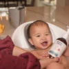 A baby in a bath seat while a parent holds the Happy Cappy medicated shampoo bottle