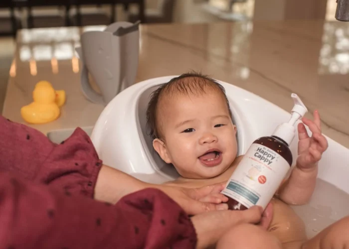 A baby in a bath seat while a parent holds the Happy Cappy medicated shampoo bottle