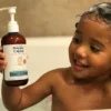 A smiling toddler in the bath holding a bottle of Happy Cappy medicated shampoo