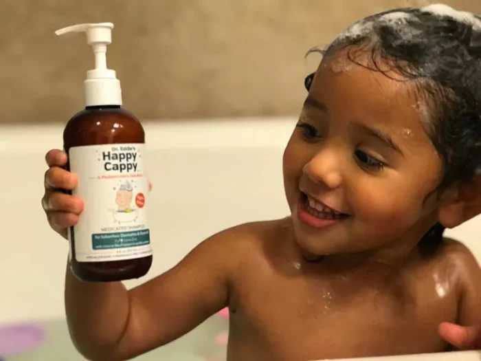 A smiling toddler in the bath holding a bottle of Happy Cappy medicated shampoo